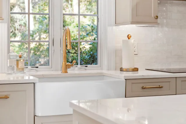 Kitchen with white farmhouse sink, gold faucet, window behind the sink, soap dispensers, paper towel holder, and stove on the right