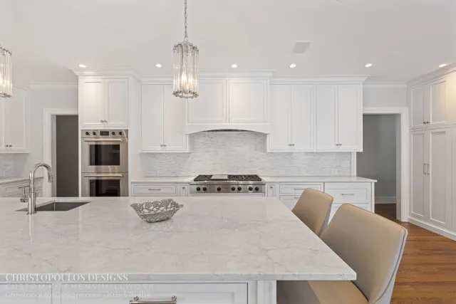 A modern white kitchen with white cabinetry, a marble island, stainless steel oven, and chandelier lighting.