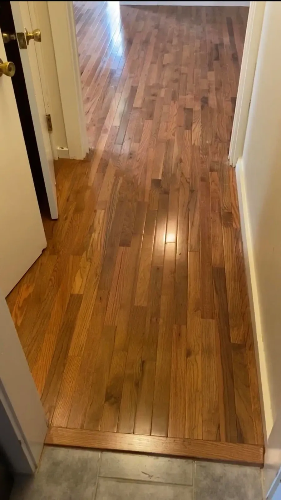 A hallway with light-brown hardwood flooring extending into an adjacent room, separated by a doorway.