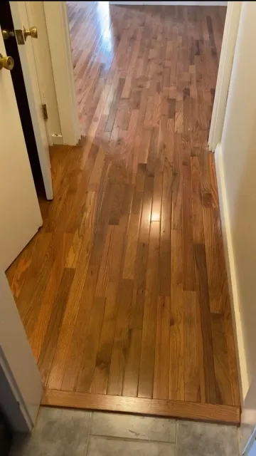A hallway with light-brown hardwood flooring extending into an adjacent room, separated by a doorway.