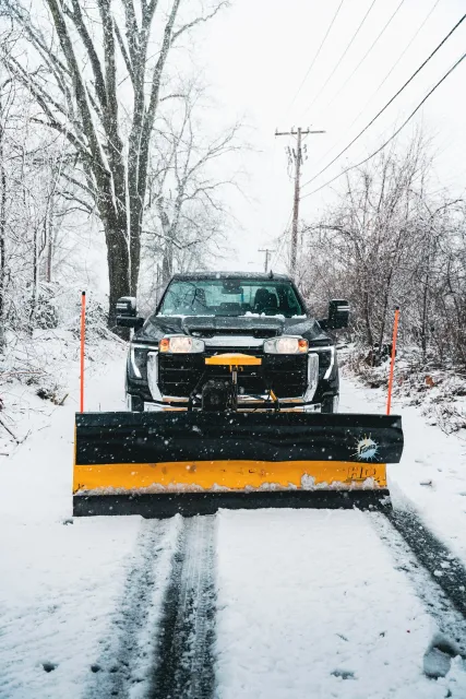 A snow-covered road with a black pickup truck in front, equipped with a yellow snowplow blade at the front, clearing snow on a winter day with snow falling and trees and power lines in the background.