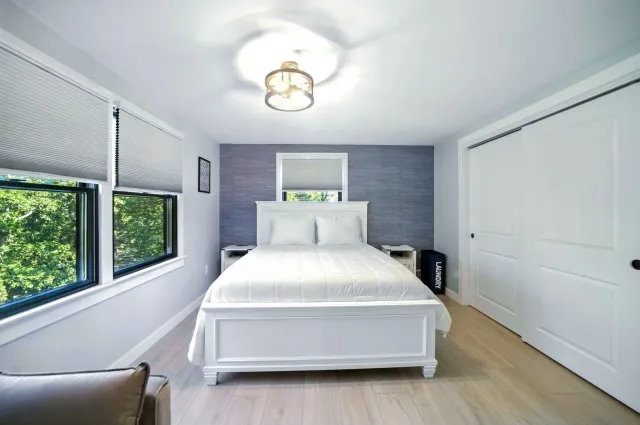Bright bedroom with white bed, closet doors, and windows with blinds on a gray-and-white wall