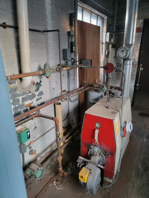 Boiler room with red industrial boiler, copper pipes, gauges, and gray ductwork against worn walls.