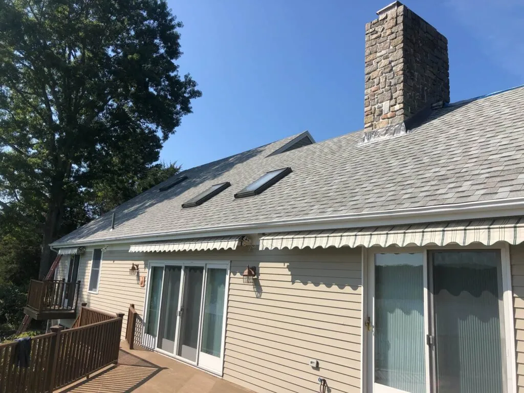 A beige house with a sloped roof featuring two skylights and a tall stone chimney. The house has a balcony with railings and two sets of sliding glass doors leading to it. Above the doors, there is a striped awning. A large tree is visible in the background.
