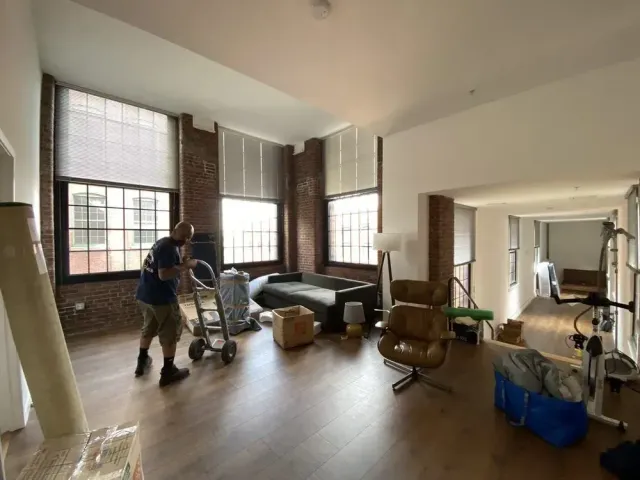 Man moves boxes on a dolly into a large, empty room with brick walls and large windows.