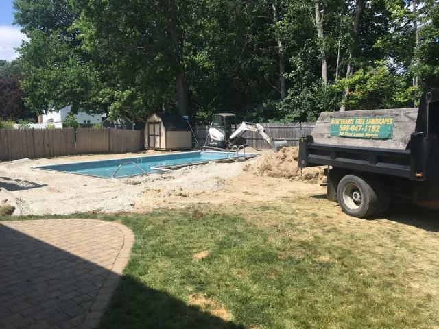 Construction work at a backyard pool with a dump truck parked on grass and a shed near a wooden fence.