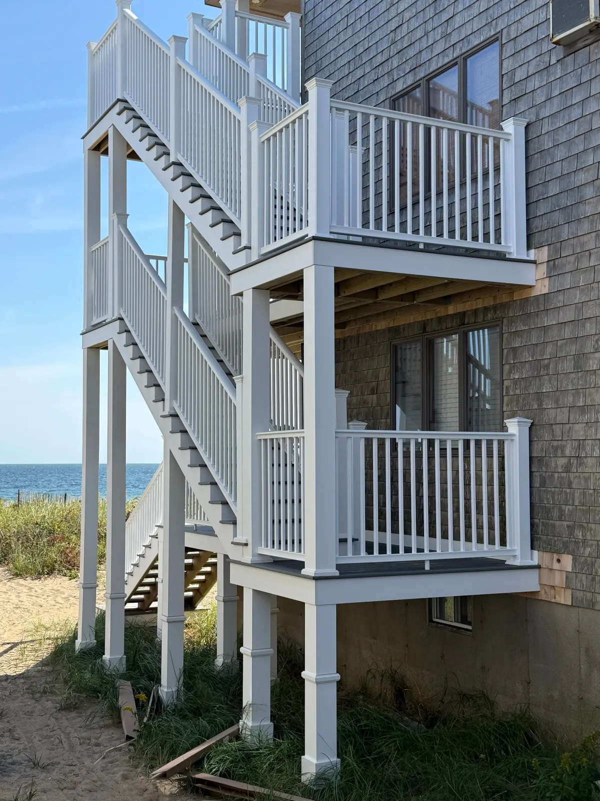 Beach house with white exterior stairs and railings leading to balconies, adjacent to a building with shingle siding, overlooking a sandy beach and ocean.