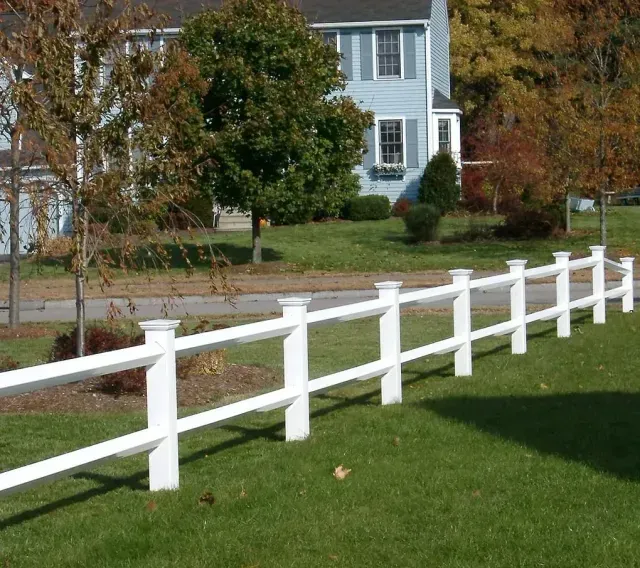 White fence along a green lawn in front of a blue house with autumn trees in the background