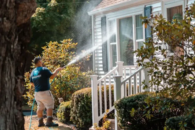 Man wearing a dark blue shirt and beige shorts power washing the white railing and siding of a house with plants around.