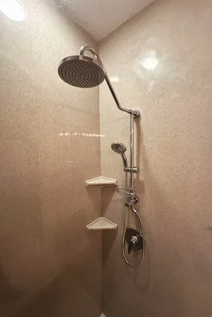 Shower with beige walls, a large overhead showerhead, a smaller handheld showerhead, a soap dish, and a water control handle.
