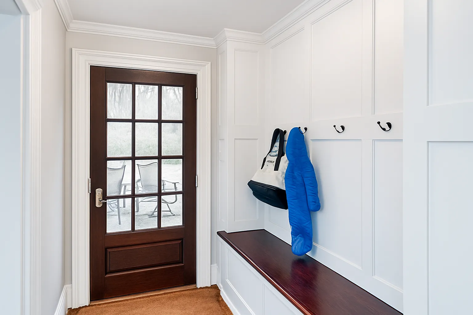 A hallway leading to a small office space with a desk, chair, and small lamp. The hallway has beige walls, a beige rug, and hanging pendant lights. A vase with orange tulips sits on a table on the right side, with a decorative wooden board on the wall nearby.