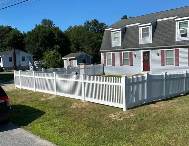 White picket fence bordering a lush green lawn, with trees and a house in the background.
