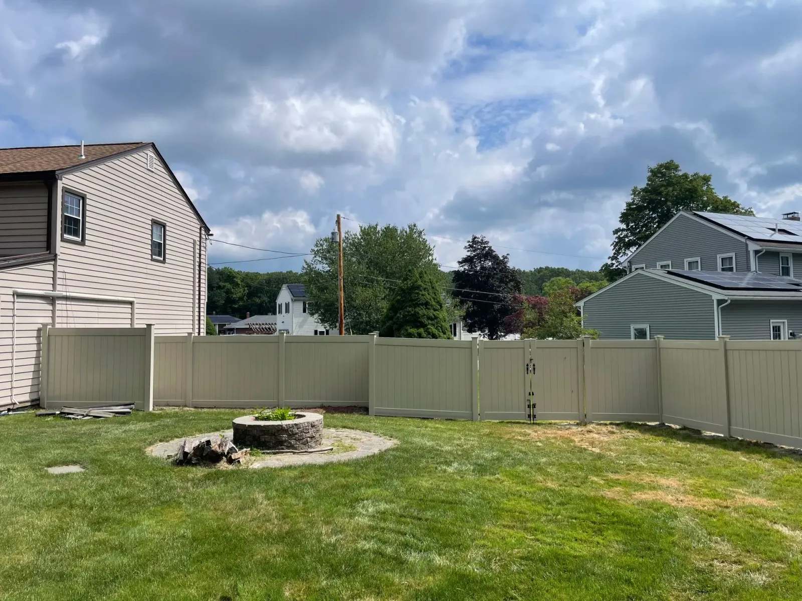 Backyard with a tan vinyl fence, fire pit, and green grass. Houses and trees in the background under a cloudy sky.