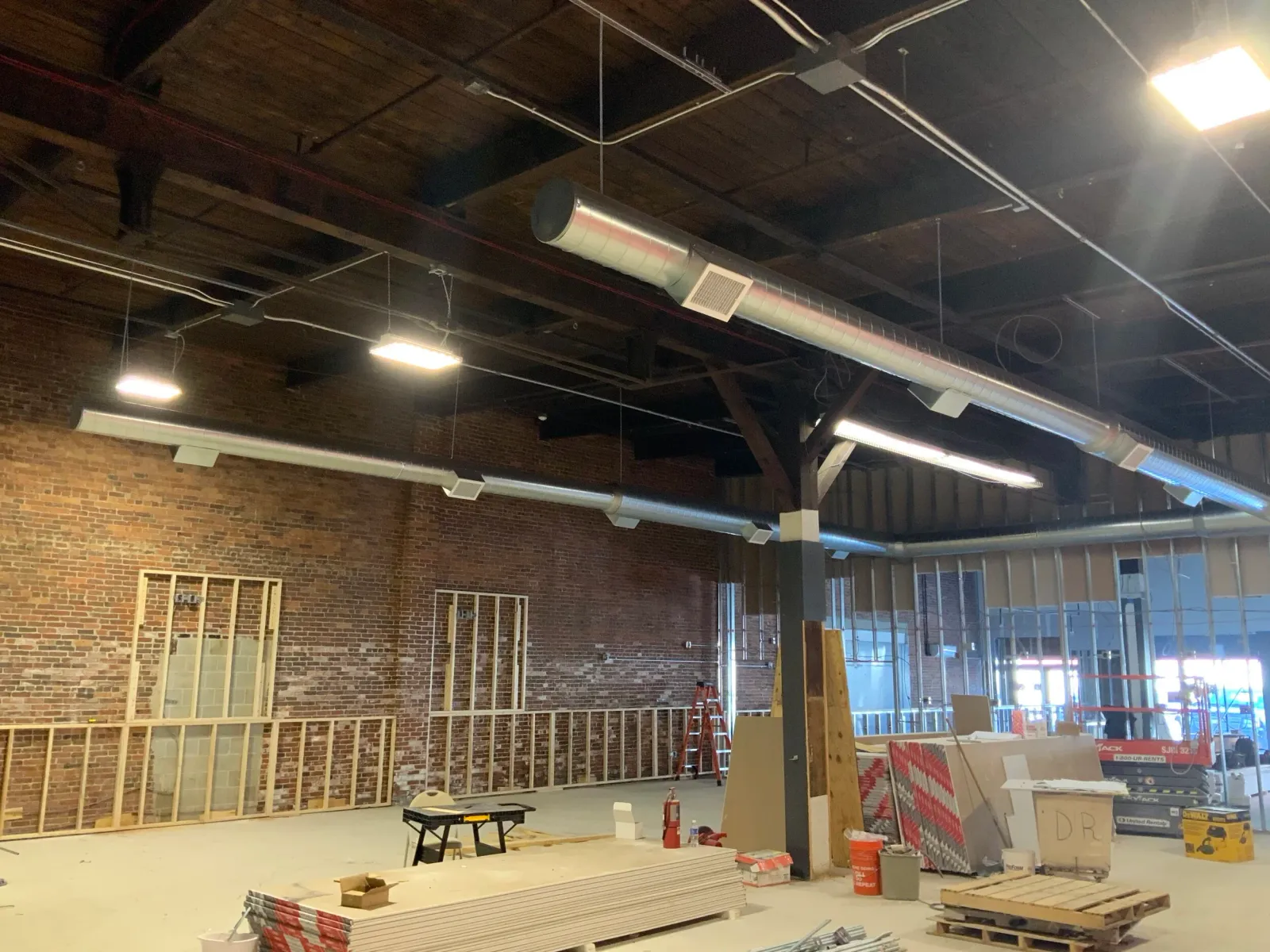 Interior of a construction site with exposed brick walls, unfinished drywall, ceiling ducts, and construction materials scattered on the floor.