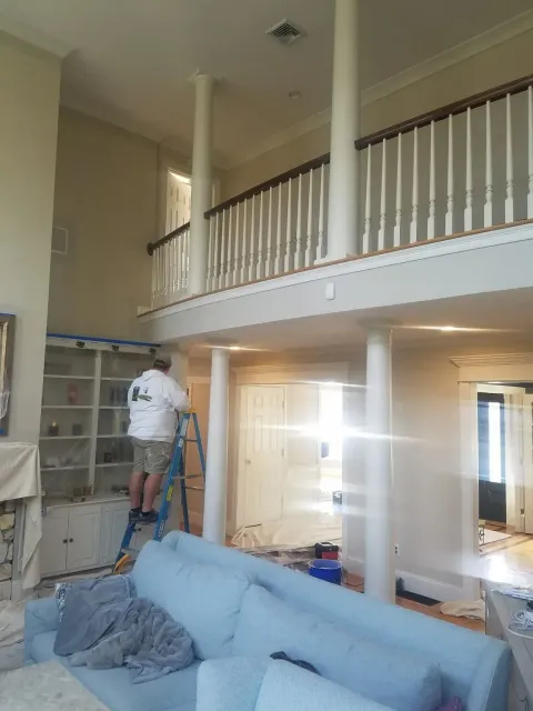 A person on a ladder paints a room with a high ceiling, columns, and a balcony.