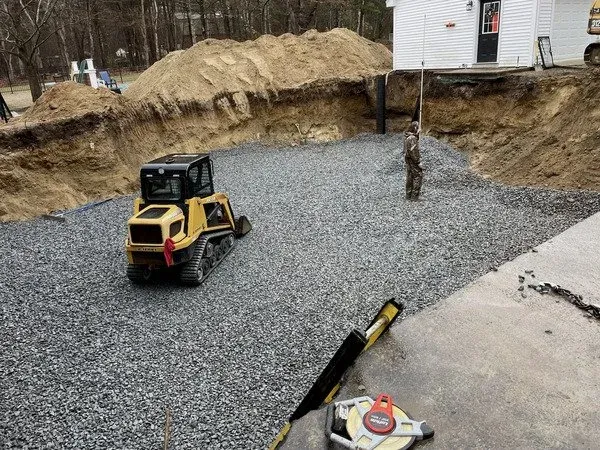 A residential construction site with a mini bulldozer, gravel, and an excavation worker in camouflage clothing standing within an excavated foundation near a house.