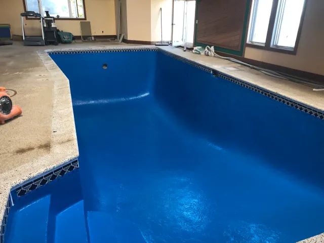 Indoor swimming pool under construction with blue waterproof coating, surrounded by a beige concrete deck, construction tools and supplies on the deck, windows and doors in the background.