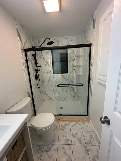Bathroom with a toilet, a white vanity, and a walk-in shower with black framing and marble walls.