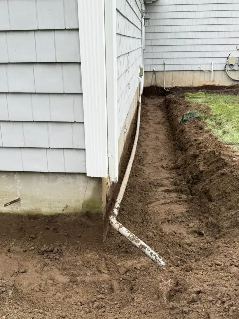 Foundation repair trench along house siding with exposed drain pipe and fresh dirt.