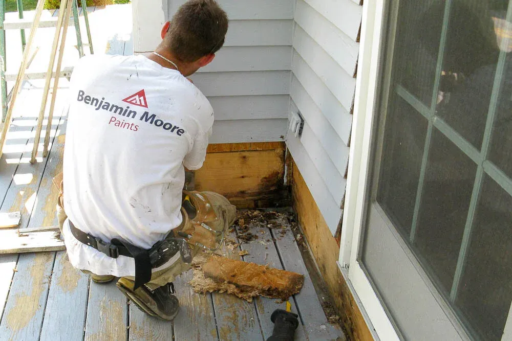 Man hunched over while repairing grey siding on the exterior of a home.