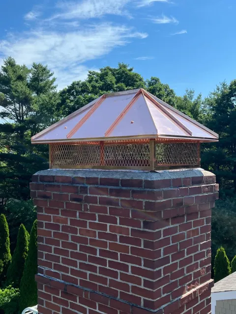 A decorative copper chimney cap with a patterned metal mesh sits atop a square red brick chimney against a blue sky.