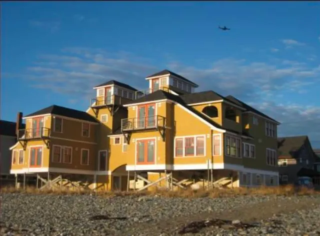 A large, multi-story beach house elevated on stilts with yellow and white siding, multiple windows, and small balconies,