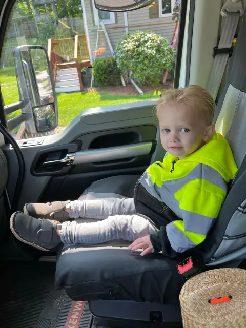 Young child in high-vis jacket sits in a truck cab, looking forward.  Sunlight streams through the window.