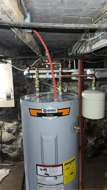 Gray water heater in a basement with exposed pipes and wiring, beneath unfinished ceiling joists.
