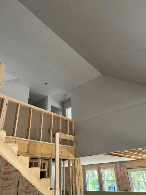 Interior of a house under construction. Drywall installed on the ceiling and walls, wooden framing visible on stairs and half-walls.