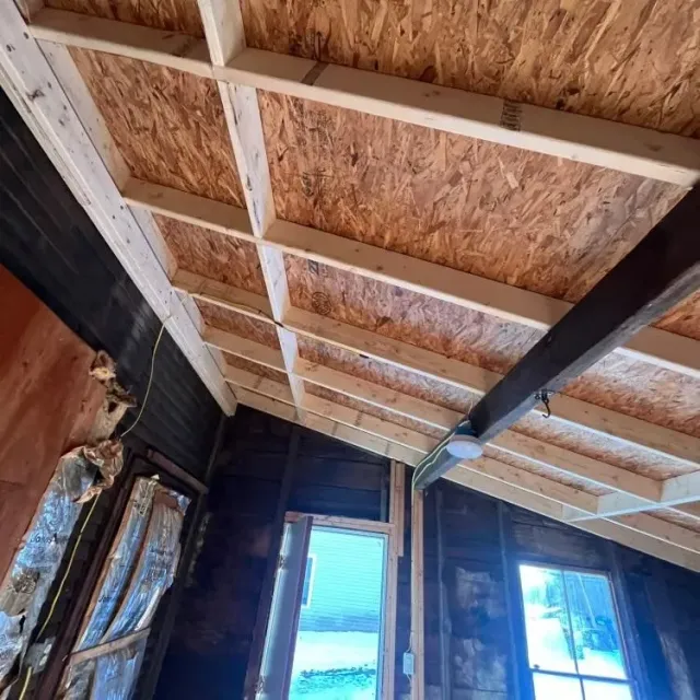 Interior ceiling under construction with exposed wooden rafters and two windows letting in daylight