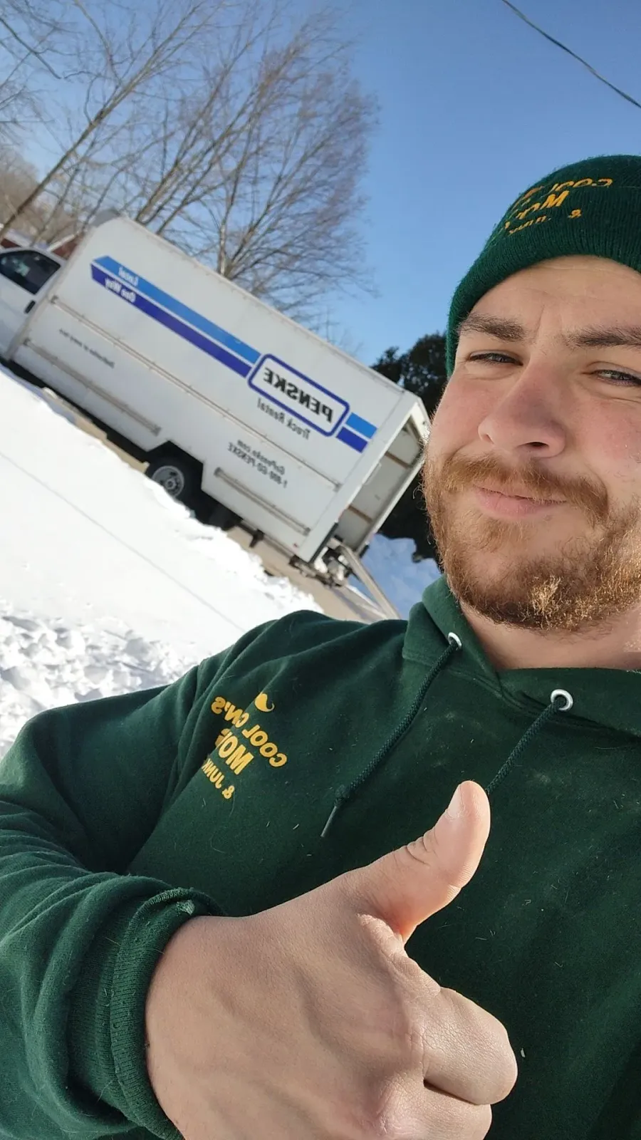 Person in green hoodie and beanie giving a thumbs-up beside a white truck in a snowy yard.