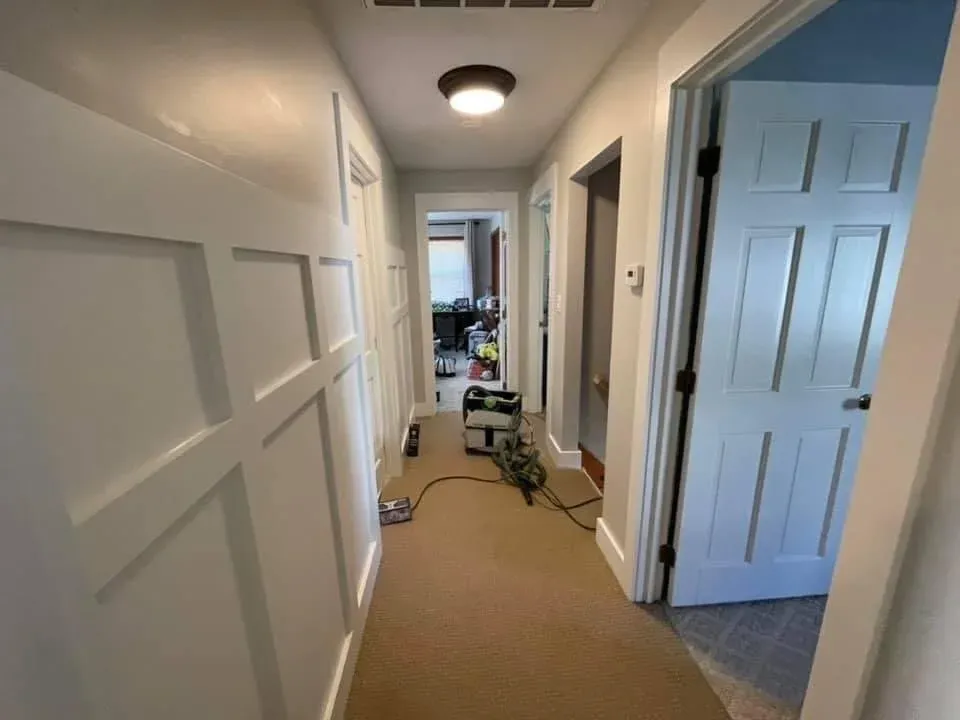 Hallway with white paneled walls, beige carpet, open doors, and cleaning equipment on the floor.