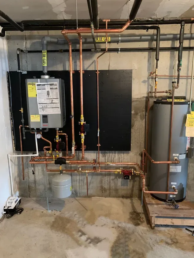 A wall-mounted tankless water heater connected to a storage tank with copper piping in a basement setting.