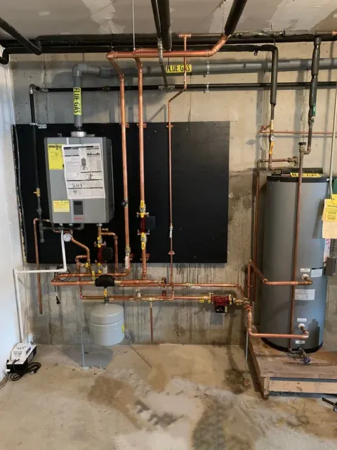 A wall-mounted tankless water heater connected to a storage tank with copper piping in a basement setting.