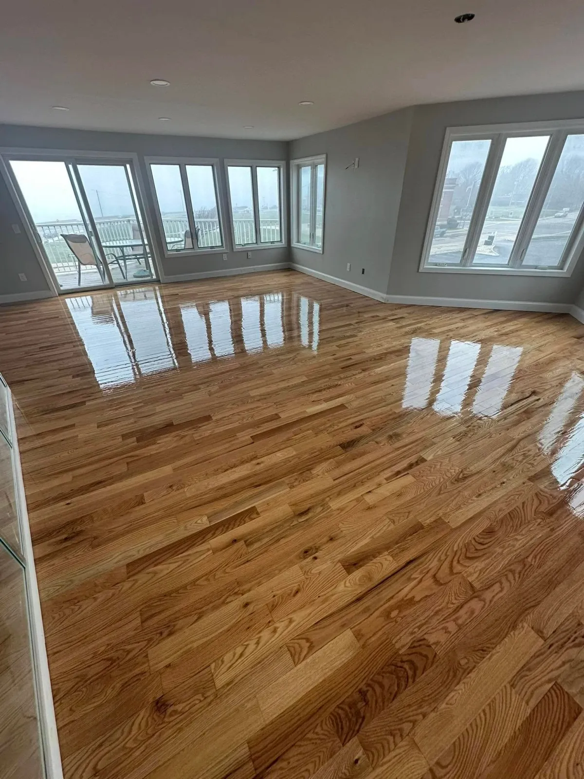 Empty living room with hardwood floors and large windows showing a balcony and outdoor view.