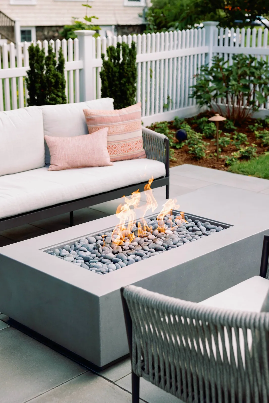 Outdoor patio with a rectangular fire pit filled with gray stones and visible flames, surrounded by a white couch with pink pillows and a woven chair, enclosed by a white picket fence with greenery and plants in the background.