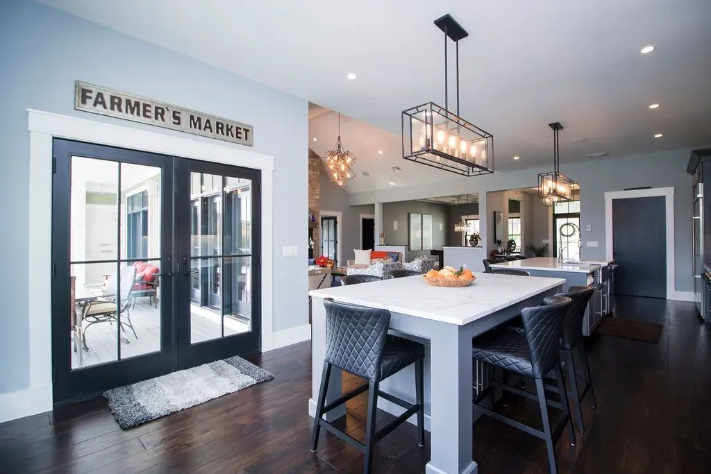 A kitchen island with gray wicker bar stools sits on a dark wood floor, next to a black-framed door with a FARMER'S MARKET sign.