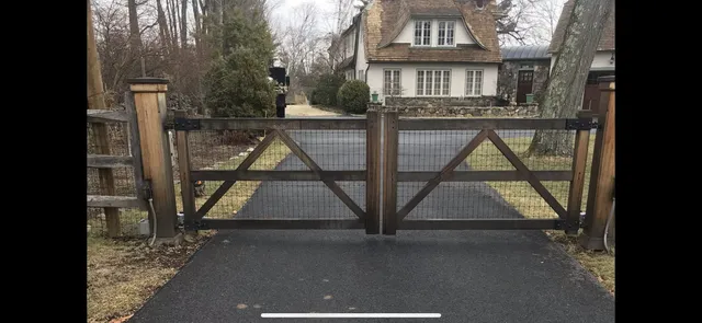 Weathered wood double driveway gate with wire mesh infill and X-bracing on paved drive with house behind