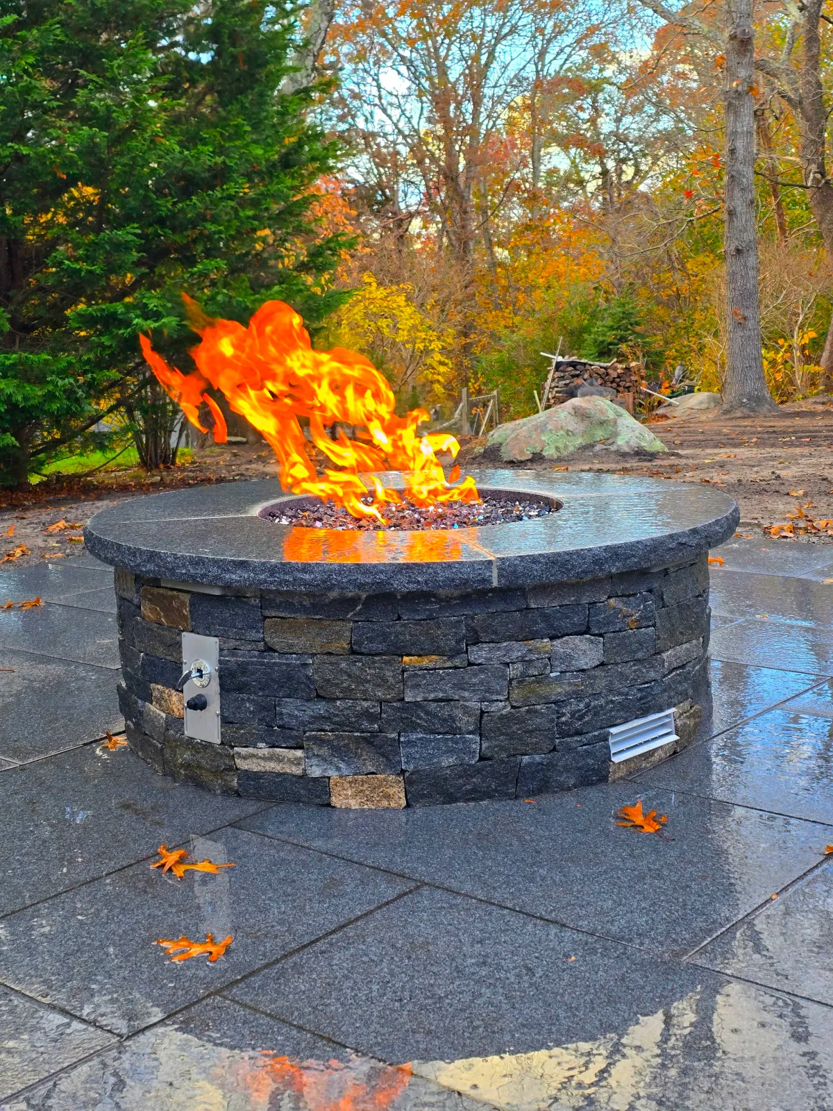 Outdoor fire pit with flames, surrounded by wet stone tiles, trees with autumn foliage, and a stack of firewood in the background.