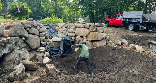 A worker in a green shirt digs into the soil in front of a newly constructed stone retaining wall near a red truck.