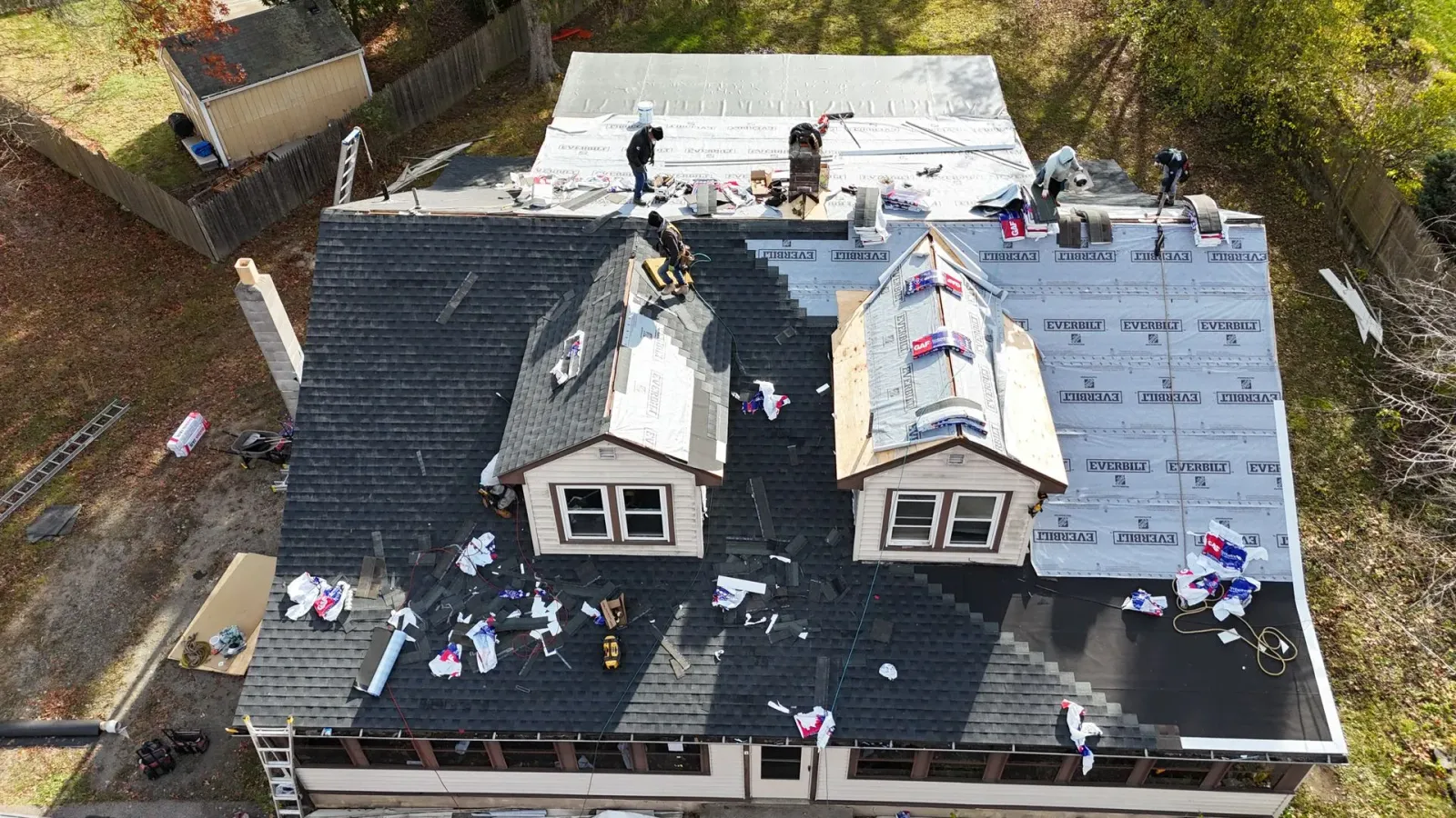 An aerial view shows multiple roofers working on a residential home's roof, replacing shingles and installing underlayment.