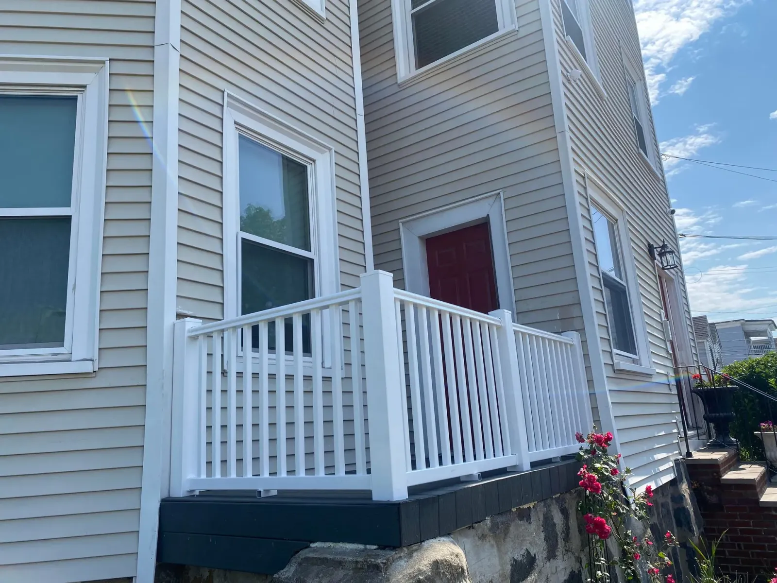 A white wooden porch railing sits in front of a tan-sided house with a red door, stone foundation, and rose bushes.