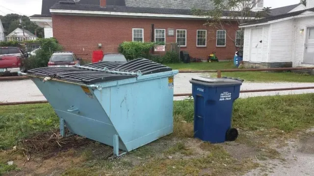 Blue dumpster and trash can on grass in front of a brick building.