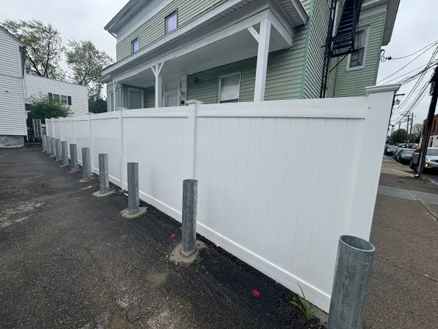 White fence with vertical metal posts lining a narrow alley next to a green building.