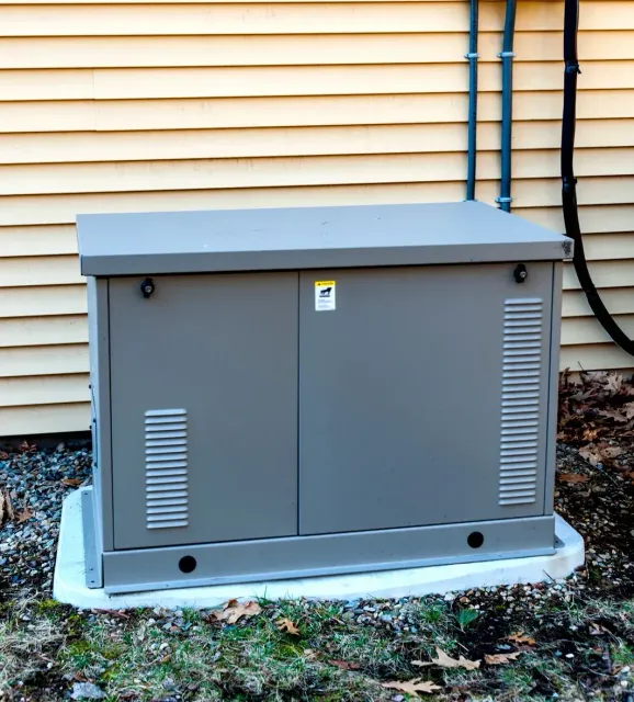 A standby generator outside a yellow house with a wood storage shed.
