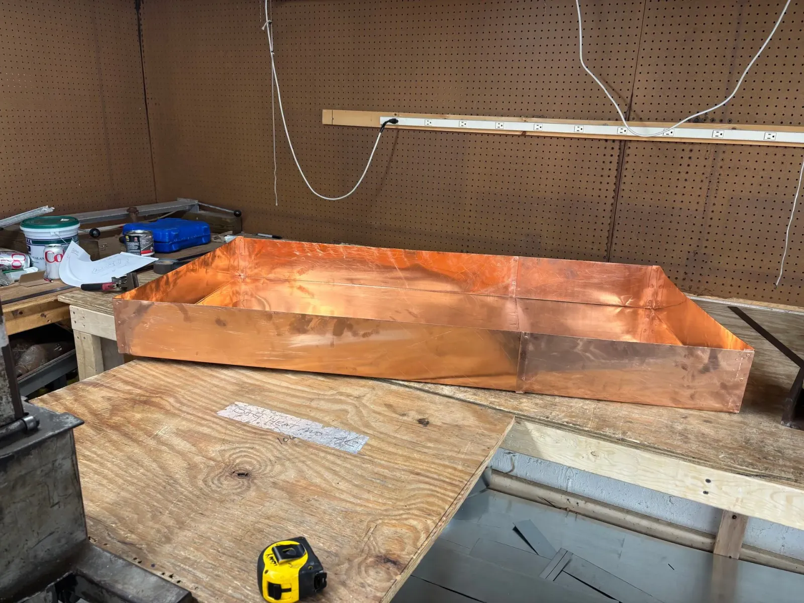 A large, rectangular copper pan sits on a wooden workbench in a workshop with a measuring tape in the foreground.