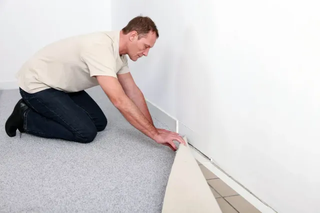 Man kneeling, installing flooring along a white wall. He is holding a floorboard.