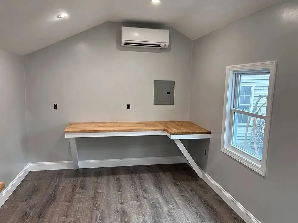 A small, light-gray room with a built-in wooden desk, window, and air conditioner.