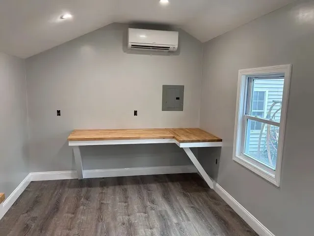 A small, light-gray room with a built-in wooden desk, window, and air conditioner.