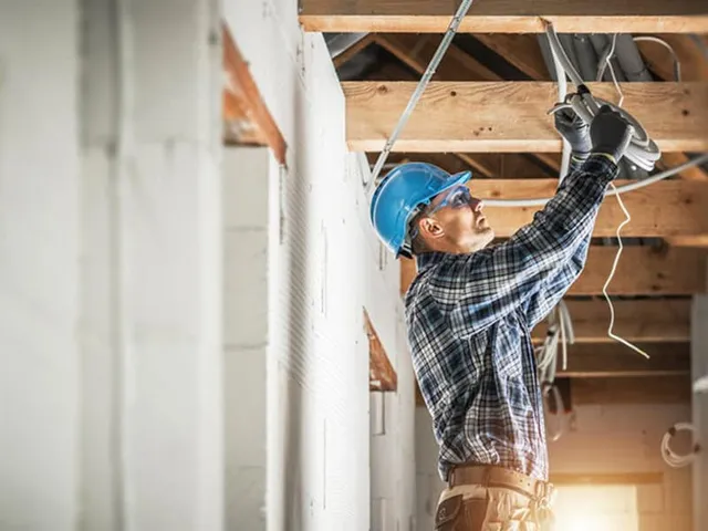 An electrician from RI wearing a blue hard hat and plaid shirt works on electrical wiring in the ceiling of a building under construction, showcasing skilled electricians Rhode Island relies on.